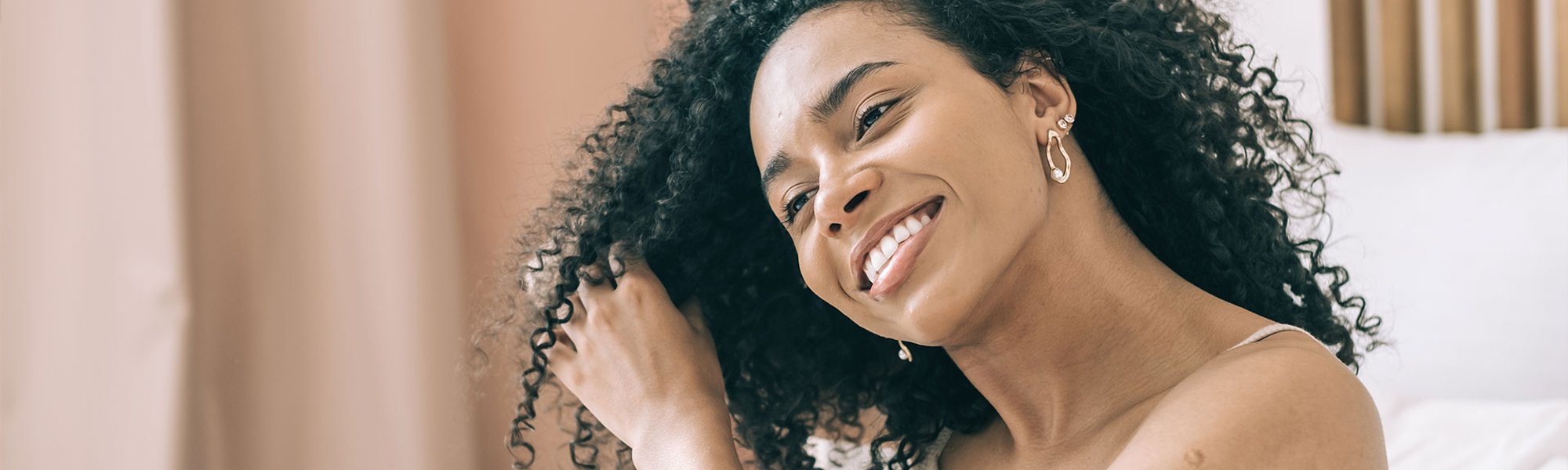 Mulher negra sorri ao tocar os cachos definidos, possivelmente com dedoliss, em seu cabelo