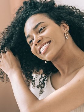 Mulher negra sorri ao tocar os cachos definidos, possivelmente com dedoliss, em seu cabelo