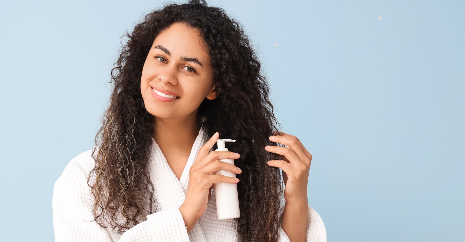Mulher sorridente aplicando definidor de cachos no seu cabelo cacheado castanho em um fundo azul.