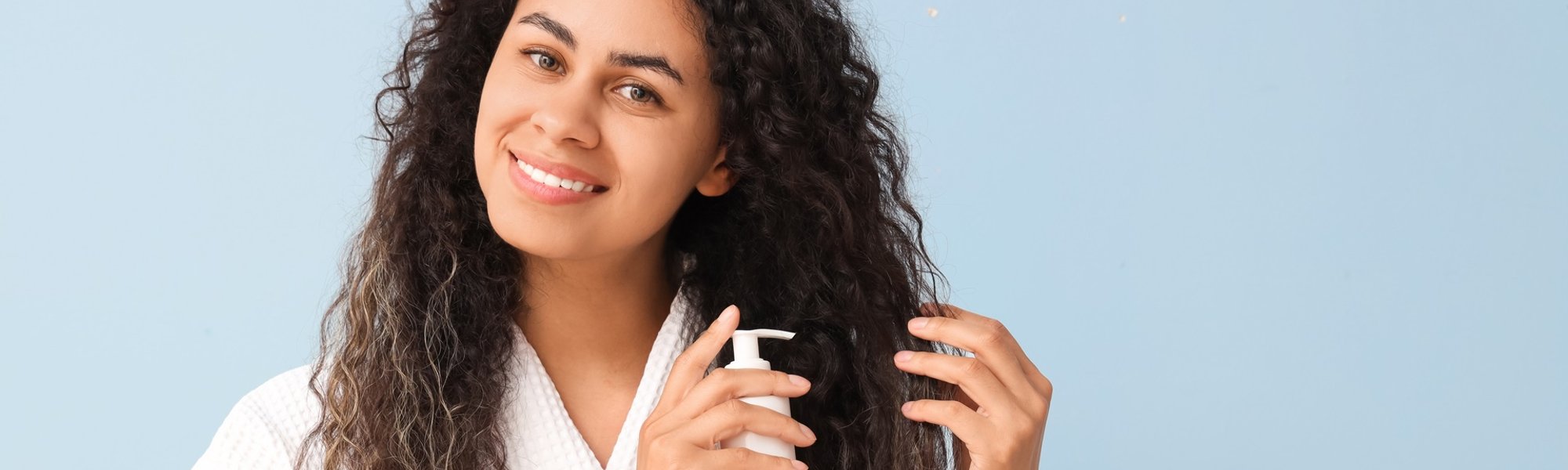 Mulher sorridente aplicando definidor de cachos no seu cabelo cacheado castanho em um fundo azul.