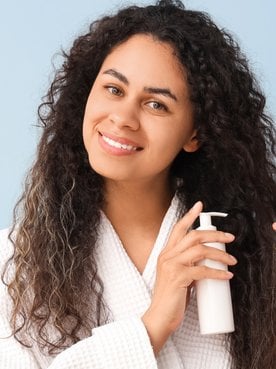 Mulher sorridente aplicando definidor de cachos no seu cabelo cacheado castanho em um fundo azul.