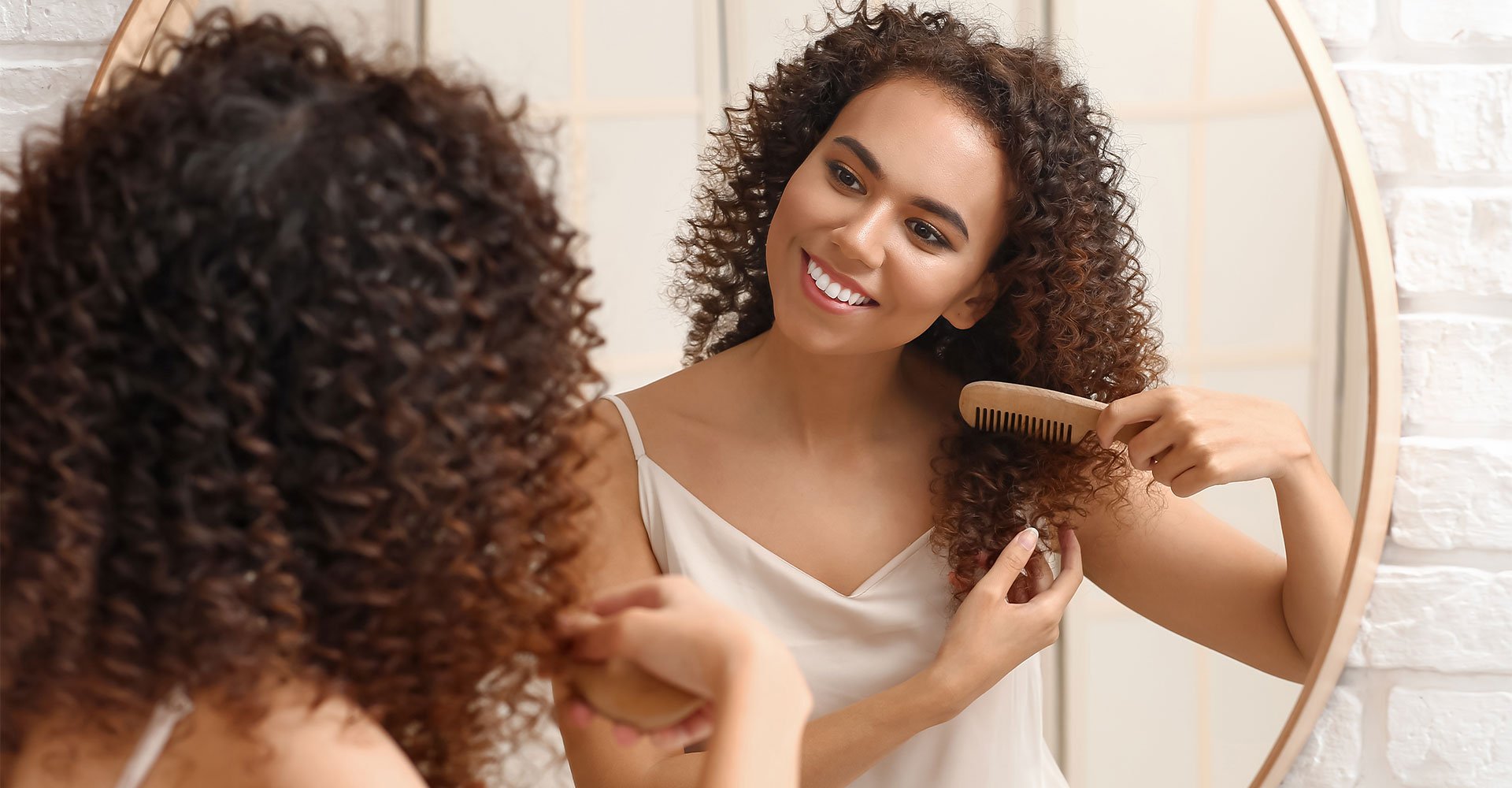 Mulher negra penteia seus cabelos nutridos com óleo de cabelo em frente ao espelho