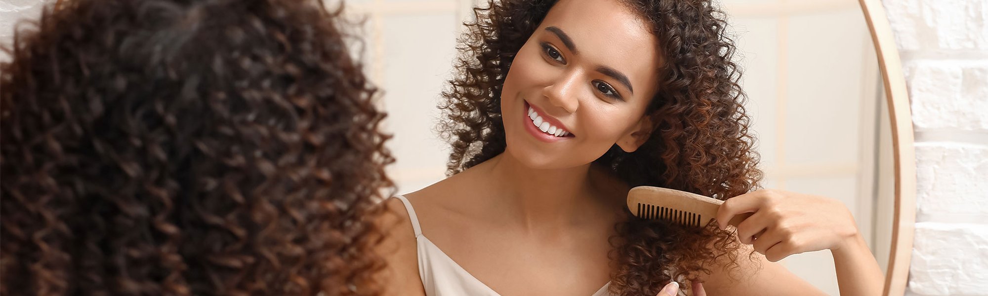 Mulher negra penteia seus cabelos nutridos com óleo de cabelo em frente ao espelho