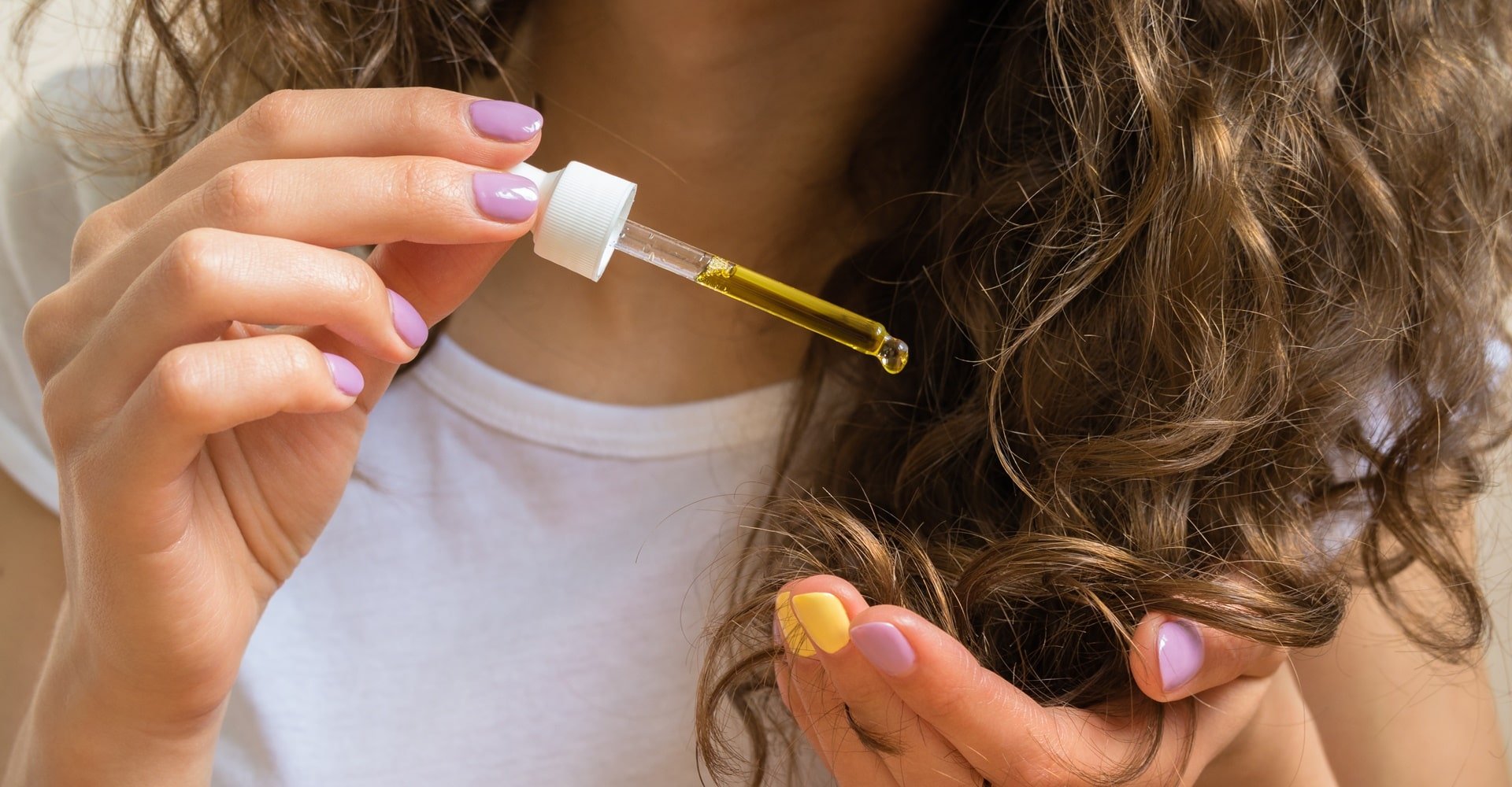 Jovem com blusa branca passando óleo de girassol com a mão em seu cabelo ondulado castanho.
