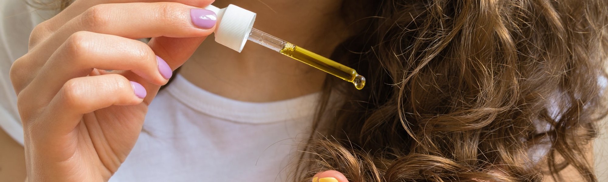 Jovem com blusa branca passando óleo de girassol com a mão em seu cabelo ondulado castanho.