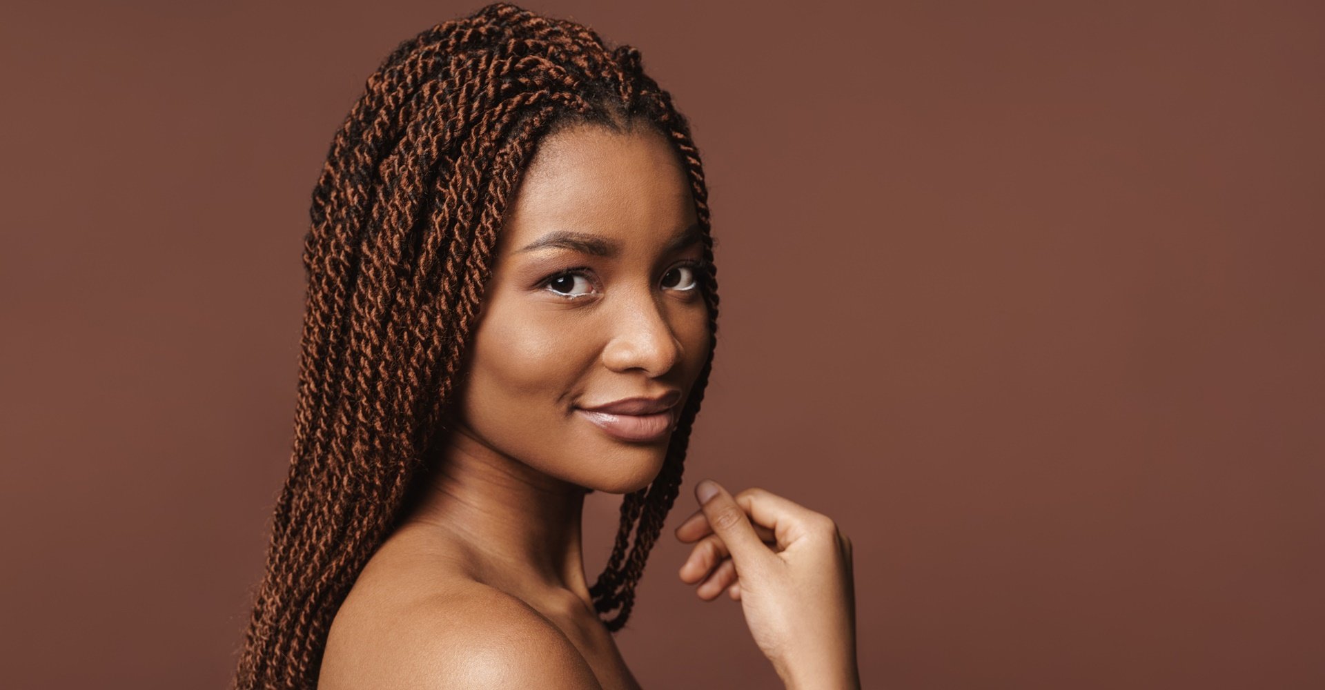 Mulher negra com trança twist no cabelo castanho olhando para a câmera em fundo marrom.