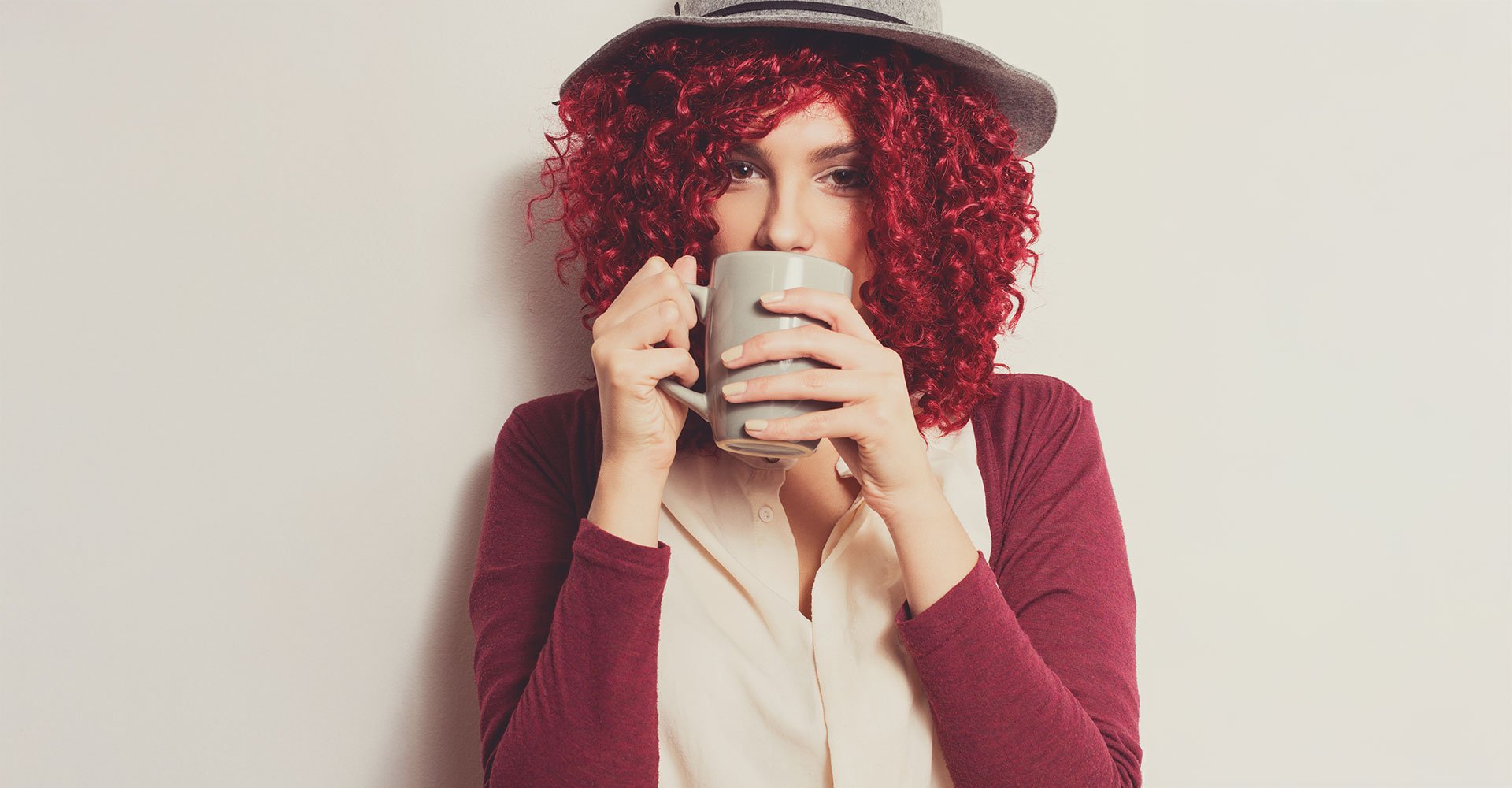 Mulher com cabelo cacheado vermelho cereja e chapéu, tomando café