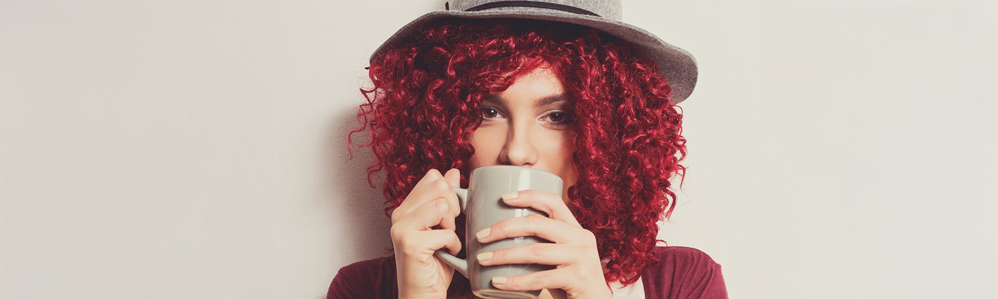 Mulher com cabelo cacheado vermelho cereja e chapéu, tomando café
