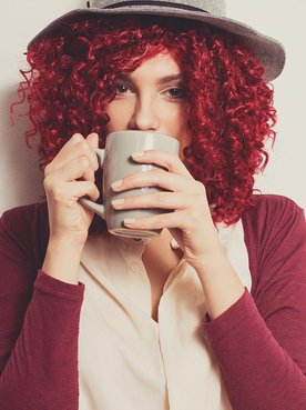 Mulher com cabelo cacheado vermelho cereja e chapéu, tomando café