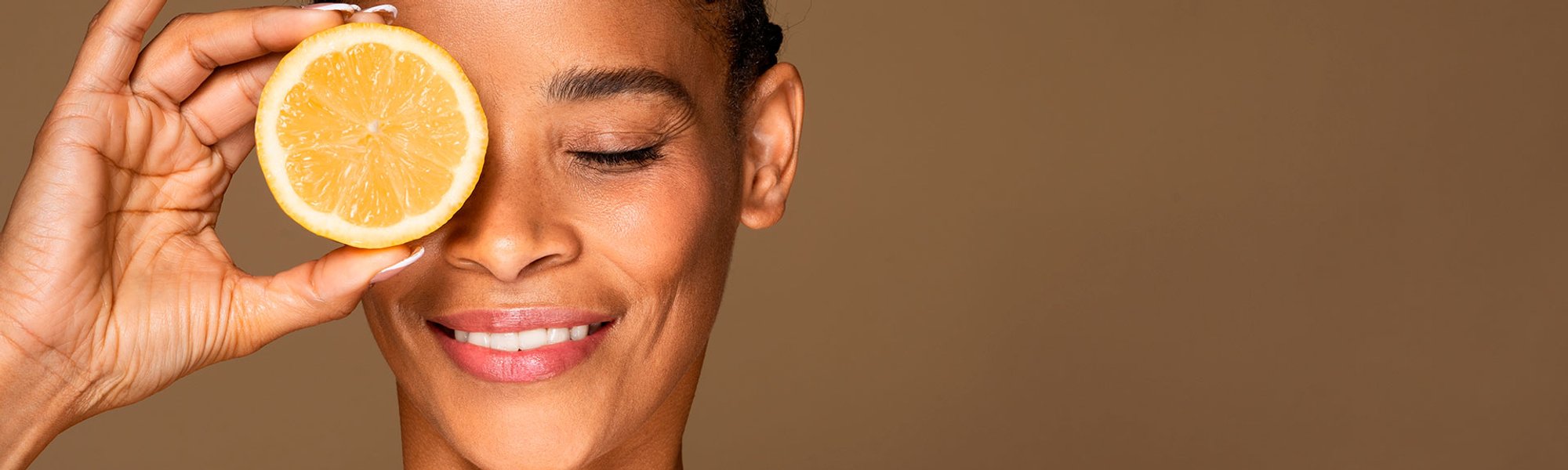 Mulher negra segurando uma laranja na altura dos olhos fazendo alusão ao uso da vitamina C no rosto