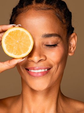 Mulher negra segurando uma laranja na altura dos olhos fazendo alusão ao uso da vitamina C no rosto