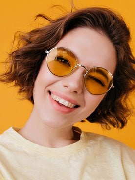 Mulher sorrindo usando óculos amarelo enquanto segura a ponta de seu cabelo curto ondulado.