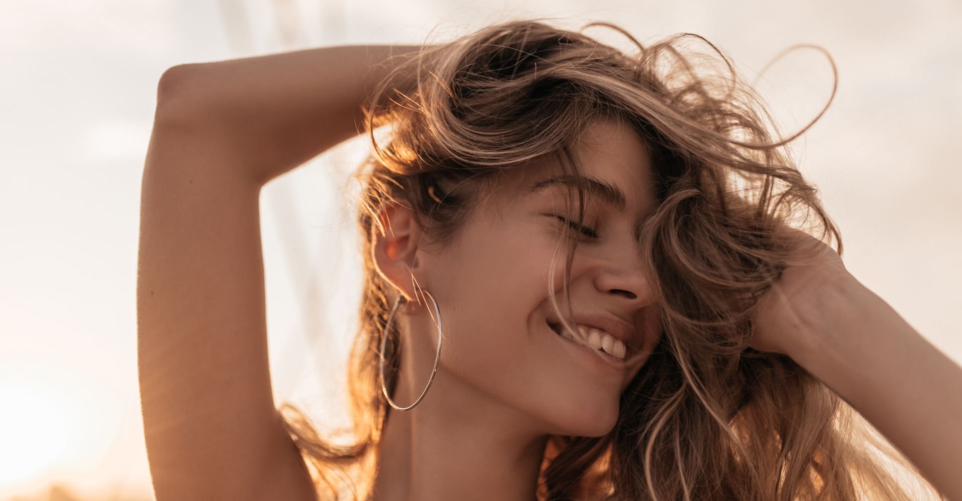 Mulher com cabelos ondulados loiro tocando os fios iluminados pela luz dourada do sol.