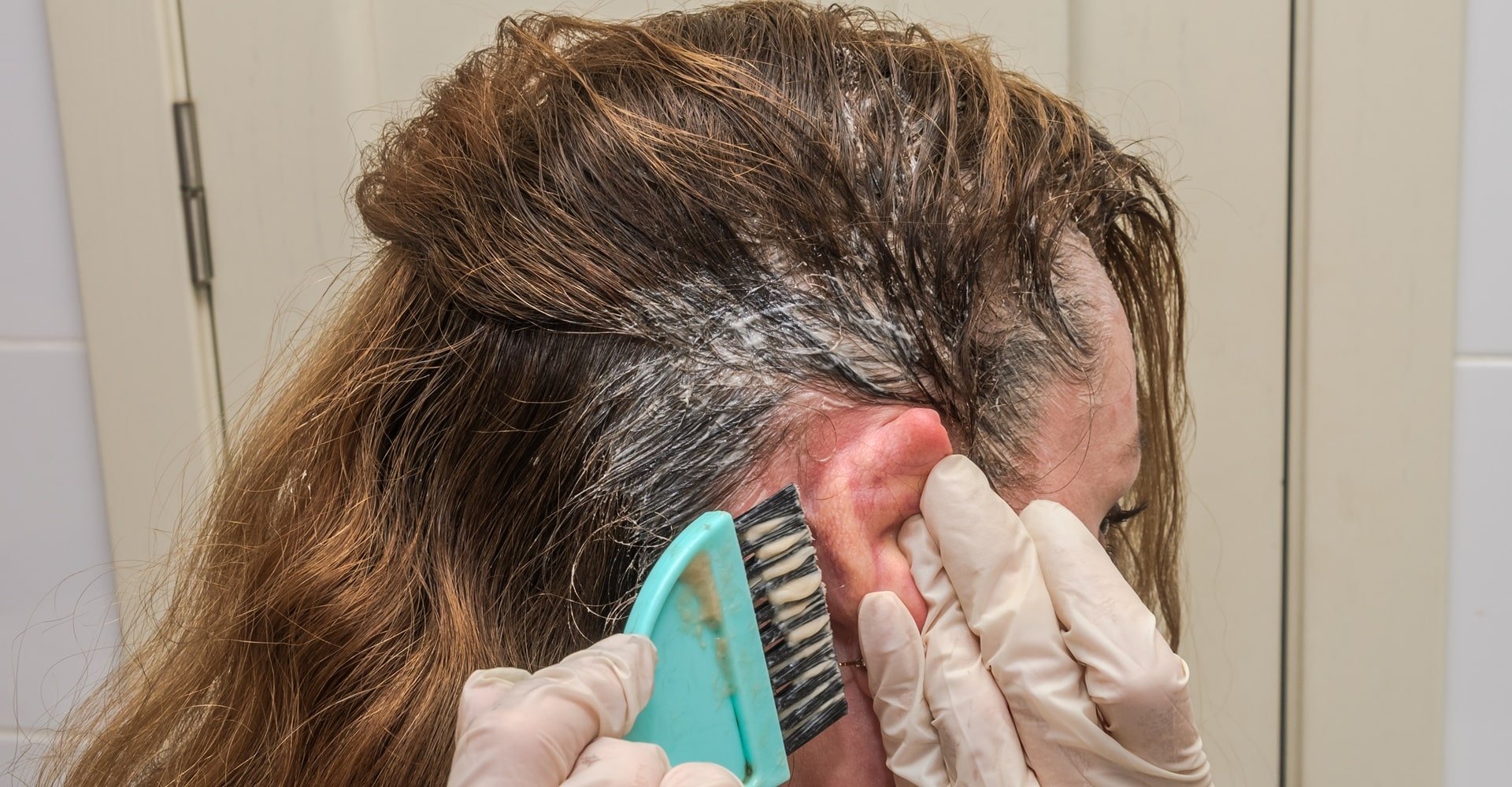Pessoa usando luvas brancas aplicando tintura de cabelo perto da orelha e linha do cabelo de alguém com cabelos castanhos