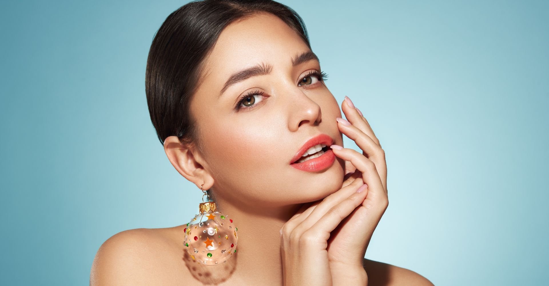 Mulher com maquiagem glamourosa, lábios pêssego e um grande brinco chamativo, posando contra um fundo azul claro. Perfeita para uma maquiagem de natal sofisticada e festiva.