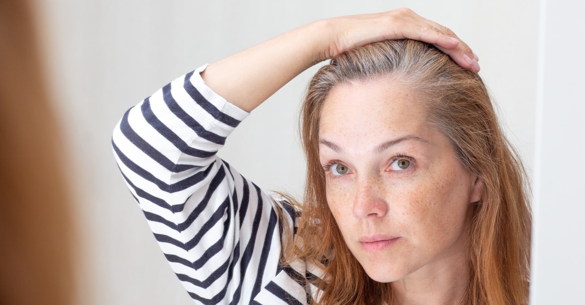 Mulher se olhando no espelho com fios de cabelo brancos.