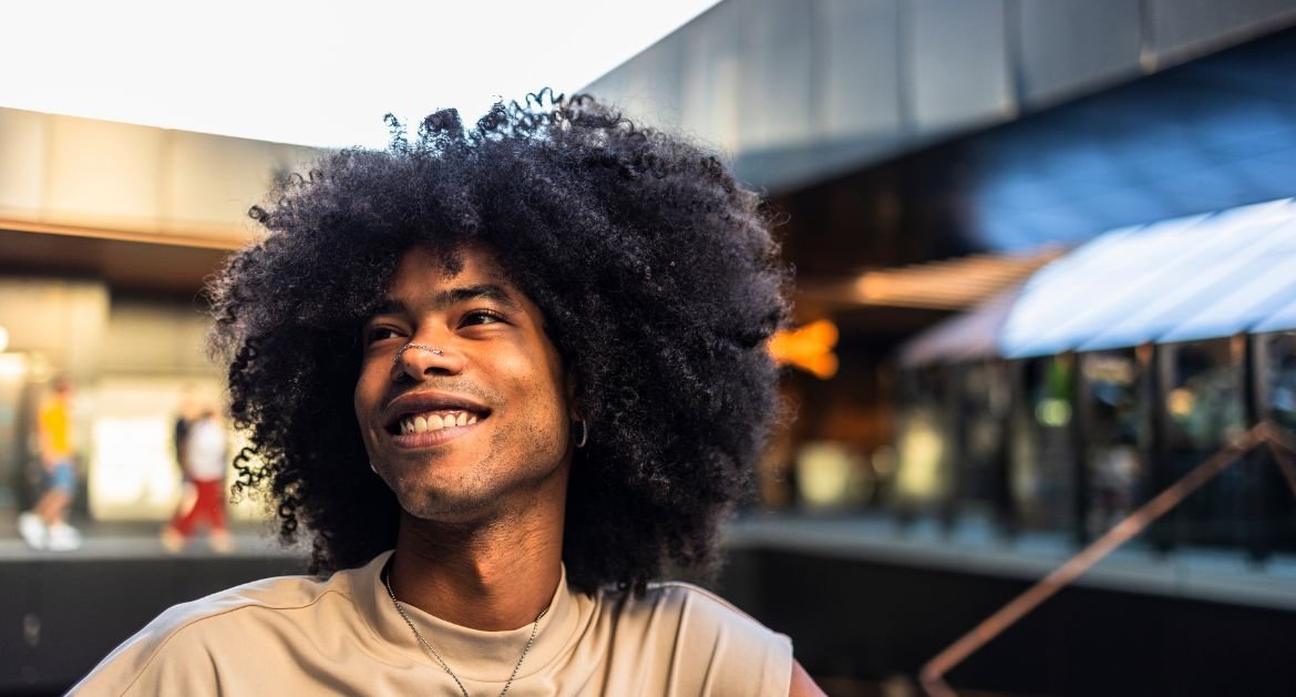 Homem com cabelo volumoso e crespo, usando camiseta bege e colar, em ambiente urbano ao ar livre.