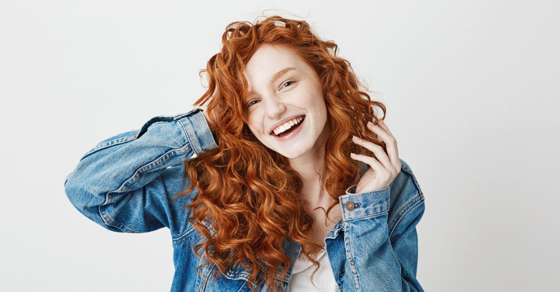 Menina sorridente com casaco jeans, blusa branca cropped e cabelo ondulado em tons de ruivo.