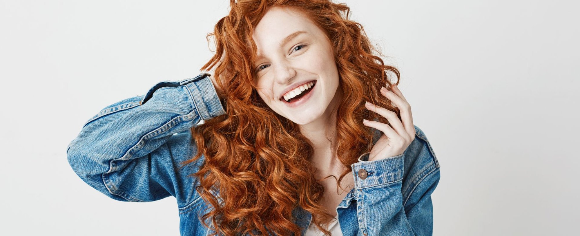 Menina sorridente com casaco jeans, blusa branca cropped e cabelo ondulado em tons de ruivo.