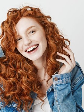 Menina sorridente com casaco jeans, blusa branca cropped e cabelo ondulado em tons de ruivo.