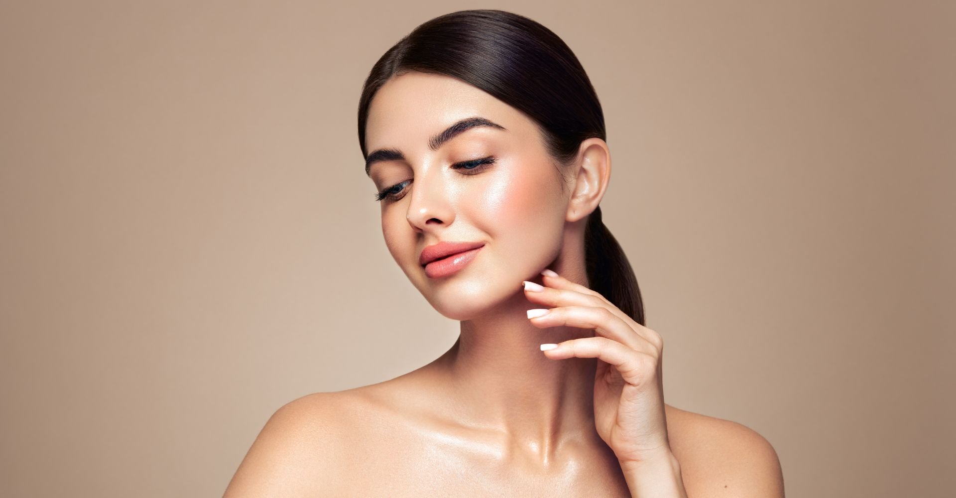 Mulher com a mão perto do rosto usando cabelo preso em rabo baixo e usando base de alta cobertura no rosto e maquiagem leve com blush levemente marcado.