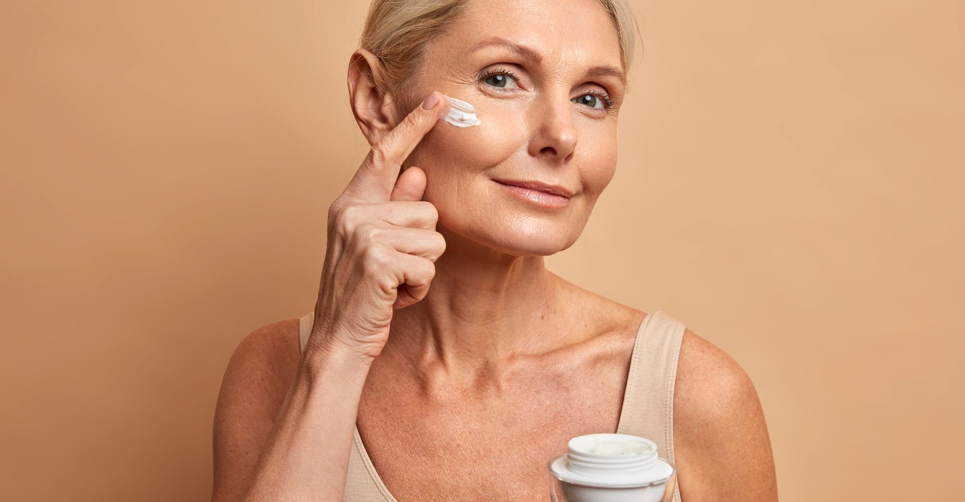 Mulher segurando pote de creme e passando em seu rosto em fundo bege.