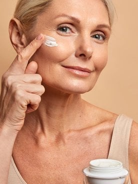 Mulher segurando pote de creme e passando em seu rosto em fundo bege.