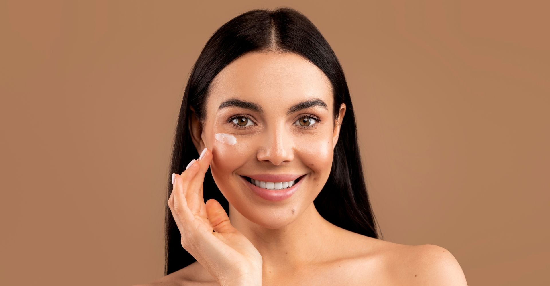 Mulher sorridente com cabelo castanho escuro passando creme para olheiras em seu rosto em fundo marrom.