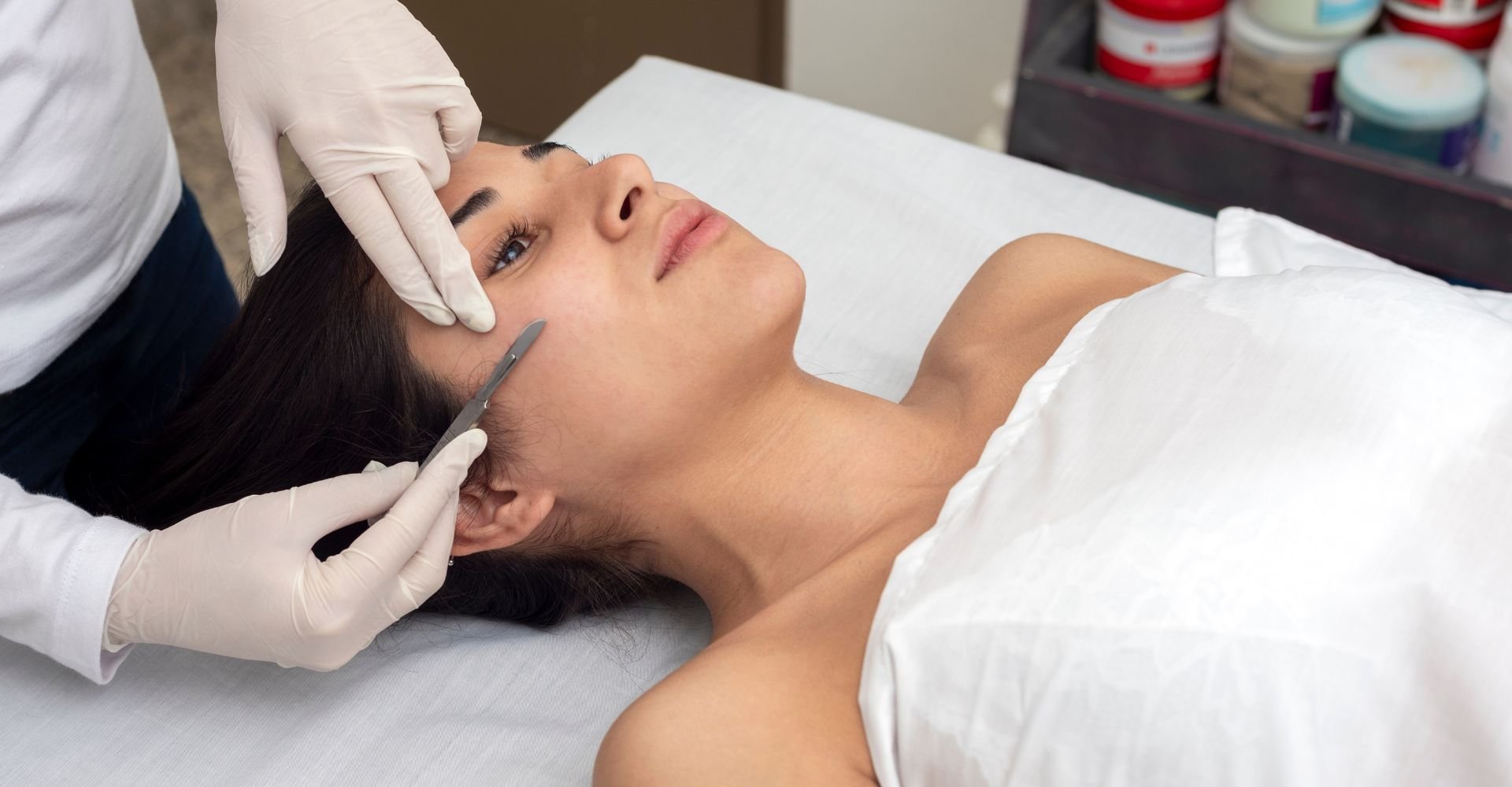 Mulher na maca de uma dermatologista realizando o procedimento de dermaplaning, que utiliza uma lâmina para esfoliar o rosto da cliente.
