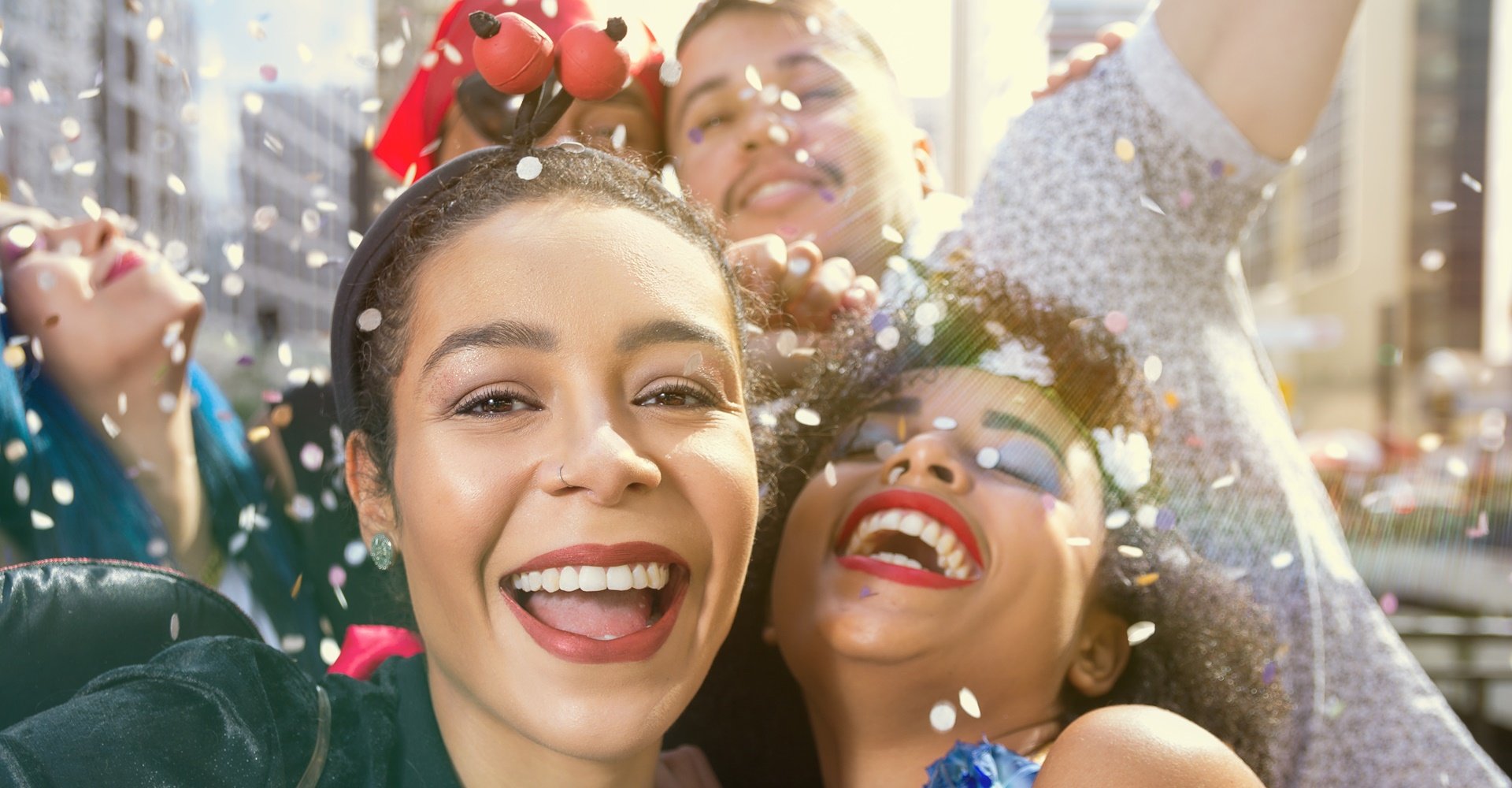 Um close-up de duas mulheres sorrindo enquanto confetes caem sobre elas. A mulher da frente usa um filtro solar com cor.