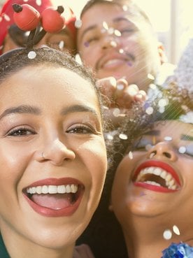 Um close-up de duas mulheres sorrindo enquanto confetes caem sobre elas. A mulher da frente usa um filtro solar com cor.