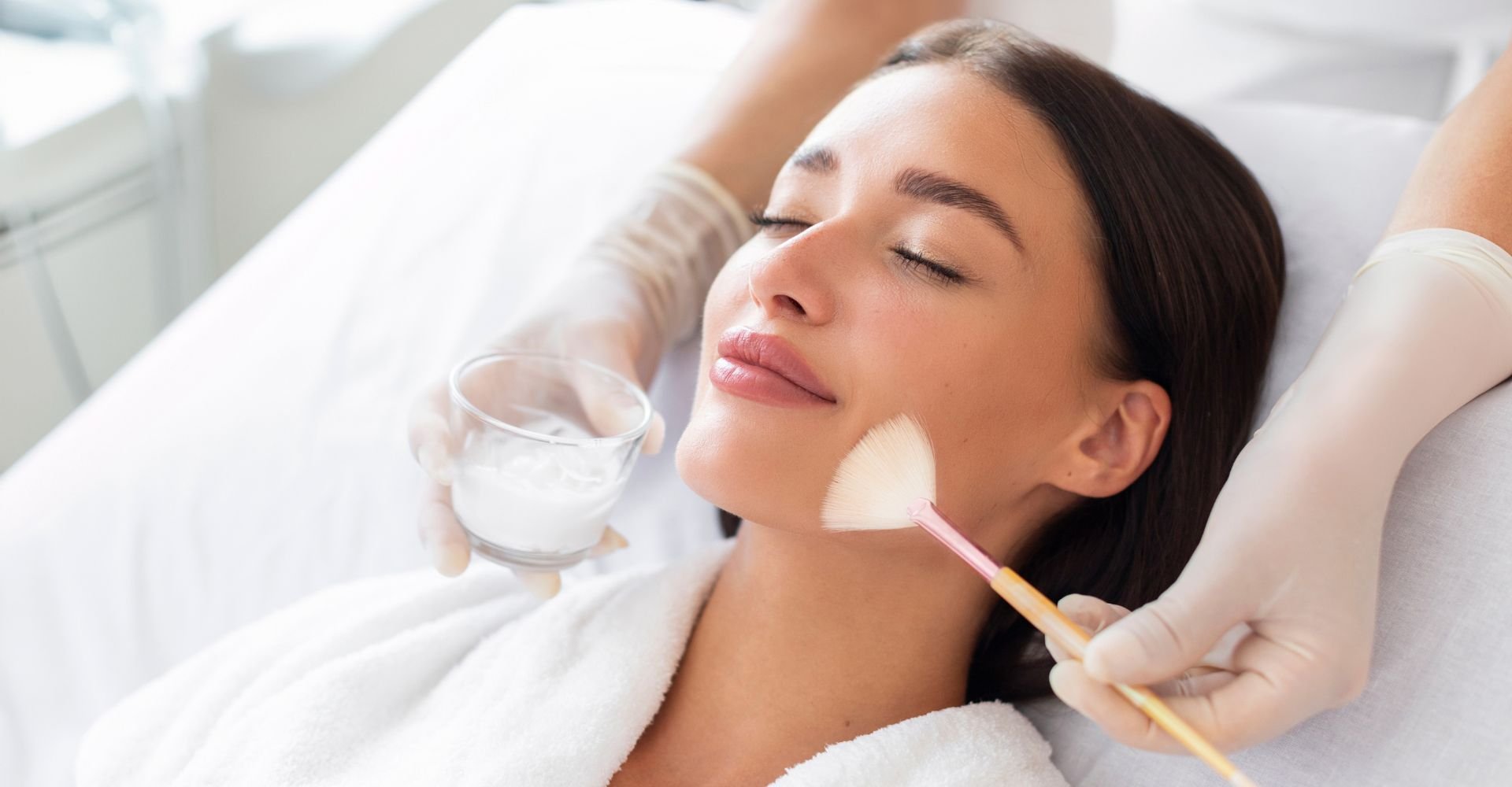 Mulher deitada em uma maca enquanto faz limpeza de pele em uma clínica, com duas mãos aplicando uma máscara facial em seu rosto.