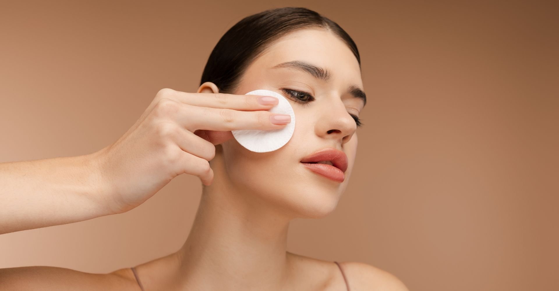 Mulher aplicando algodão com removedor de maquiagem no rosto em fundo bege.