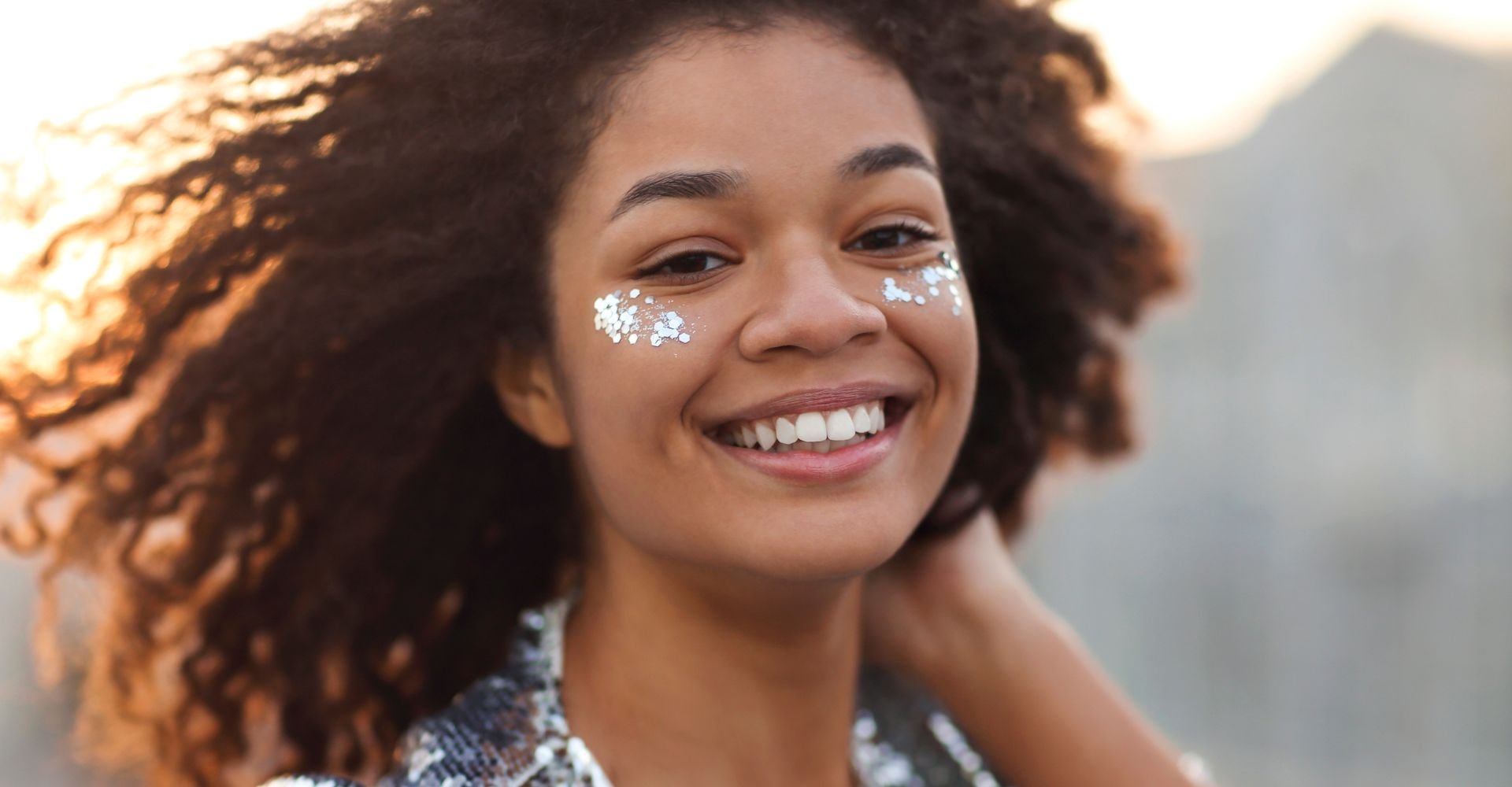 Close-up de uma mulher sorridente com cabelo crespo volumoso e natural, com glitter prateado aplicado sob os olhos. 