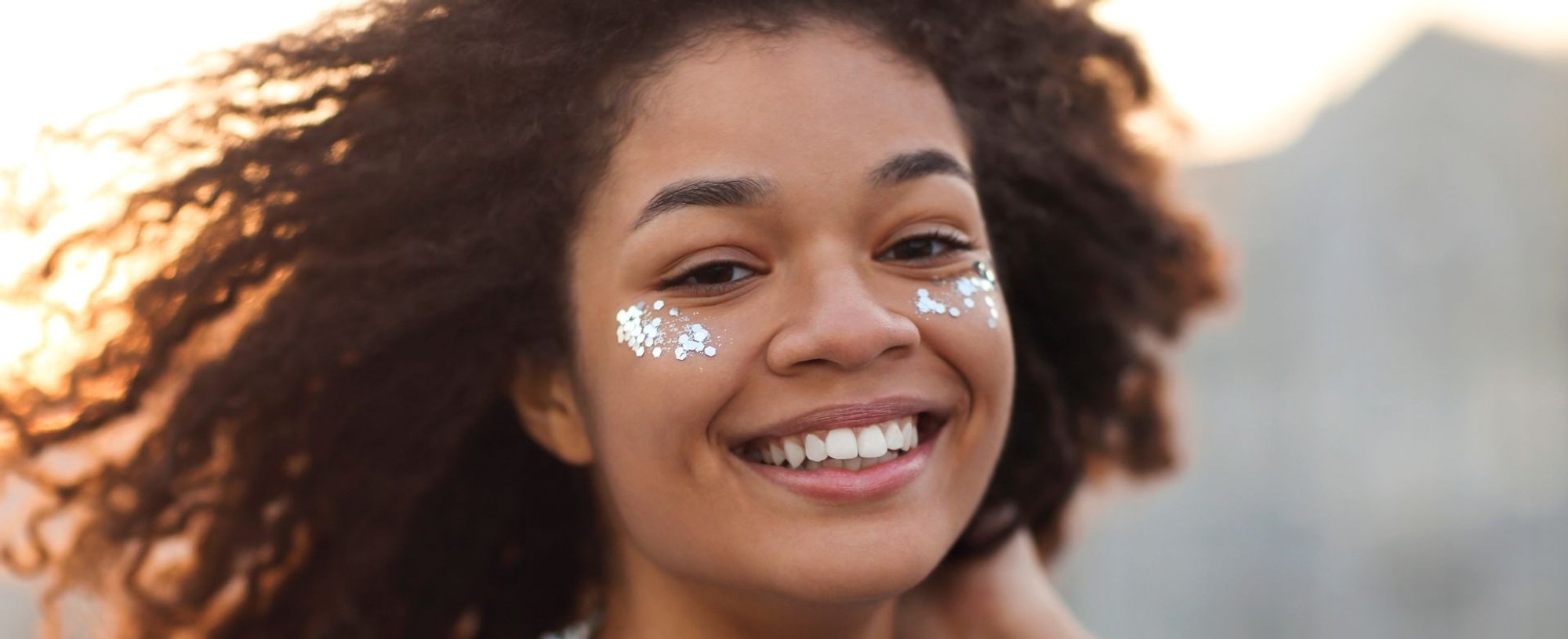 Close-up de uma mulher sorridente com cabelo crespo volumoso e natural, com glitter prateado aplicado sob os olhos.