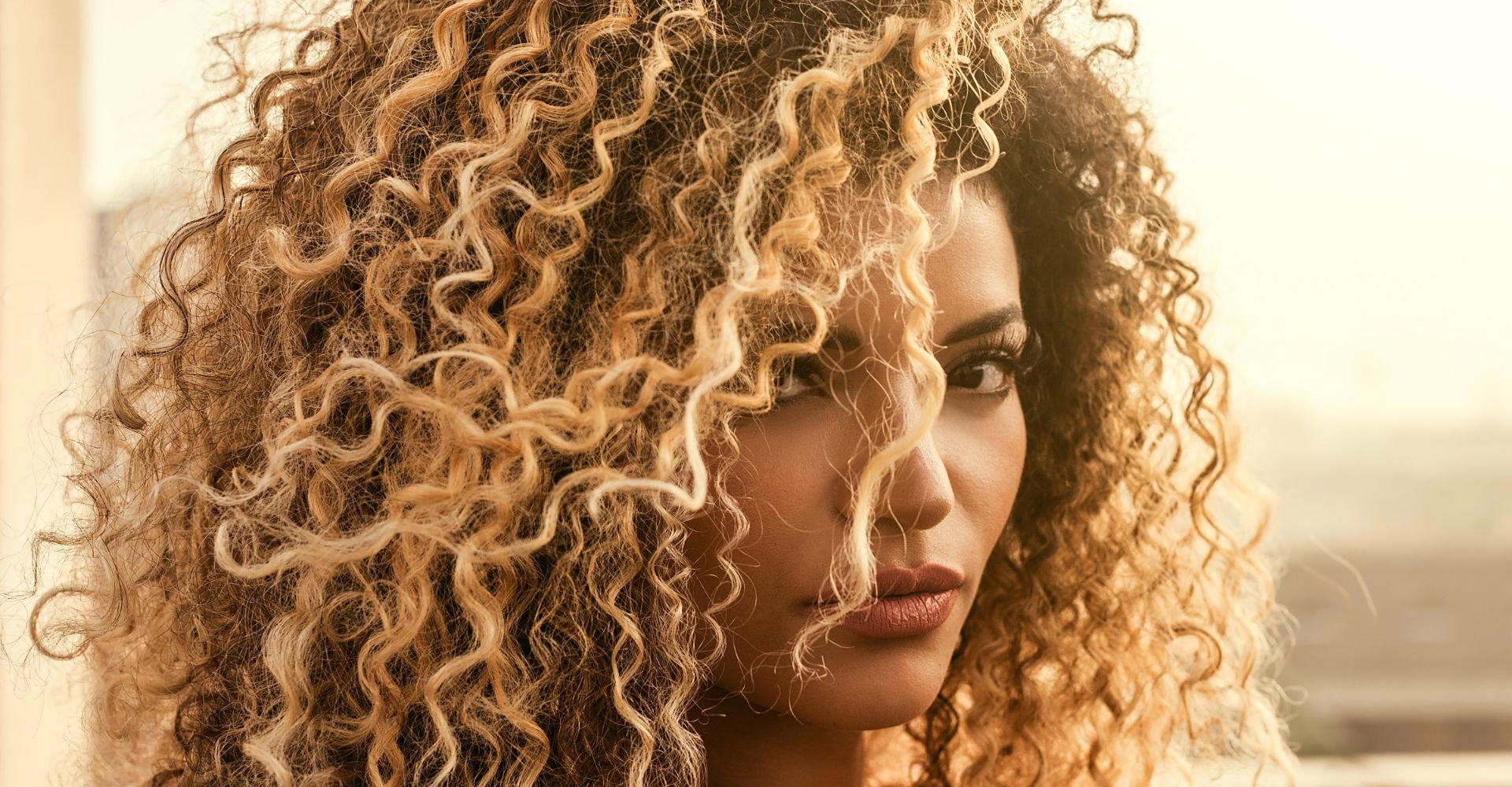Mulher com cabelos loiros, cacheados e volumosos contra o sol.