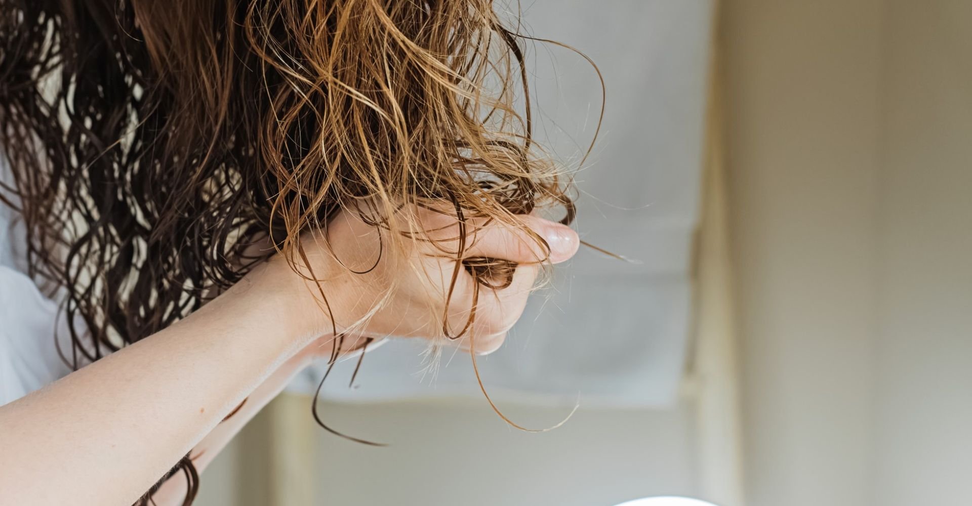 Mulher com cabelos cacheados e úmidos observa as pontas dos fios diante do espelho após lavar o cabelo, etapa ideal para aplicar máscara capilar.