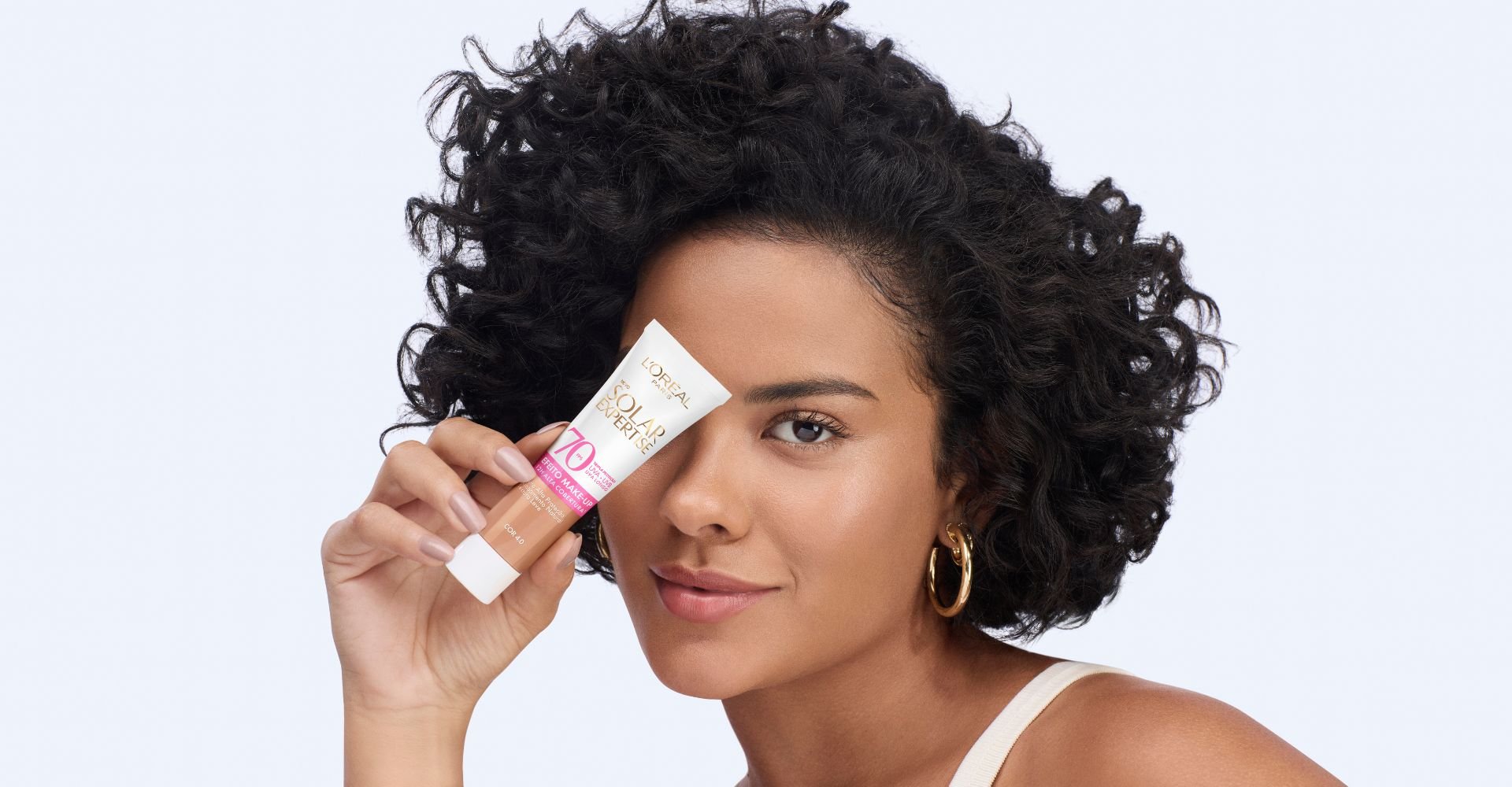  Bella Campos apresentando o protetor solar com cor L'Oréal Paris Solar Expertise Efeito Make-Up FPS 70, segurando o tubo na altura dos olhos e com um sorriso.