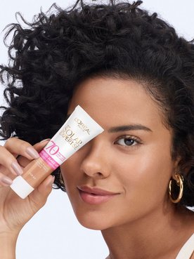 Bella Campos apresentando o protetor solar com cor L'Oréal Paris Solar Expertise Efeito Make-Up FPS 70, segurando o tubo na altura dos olhos e com um sorriso.
