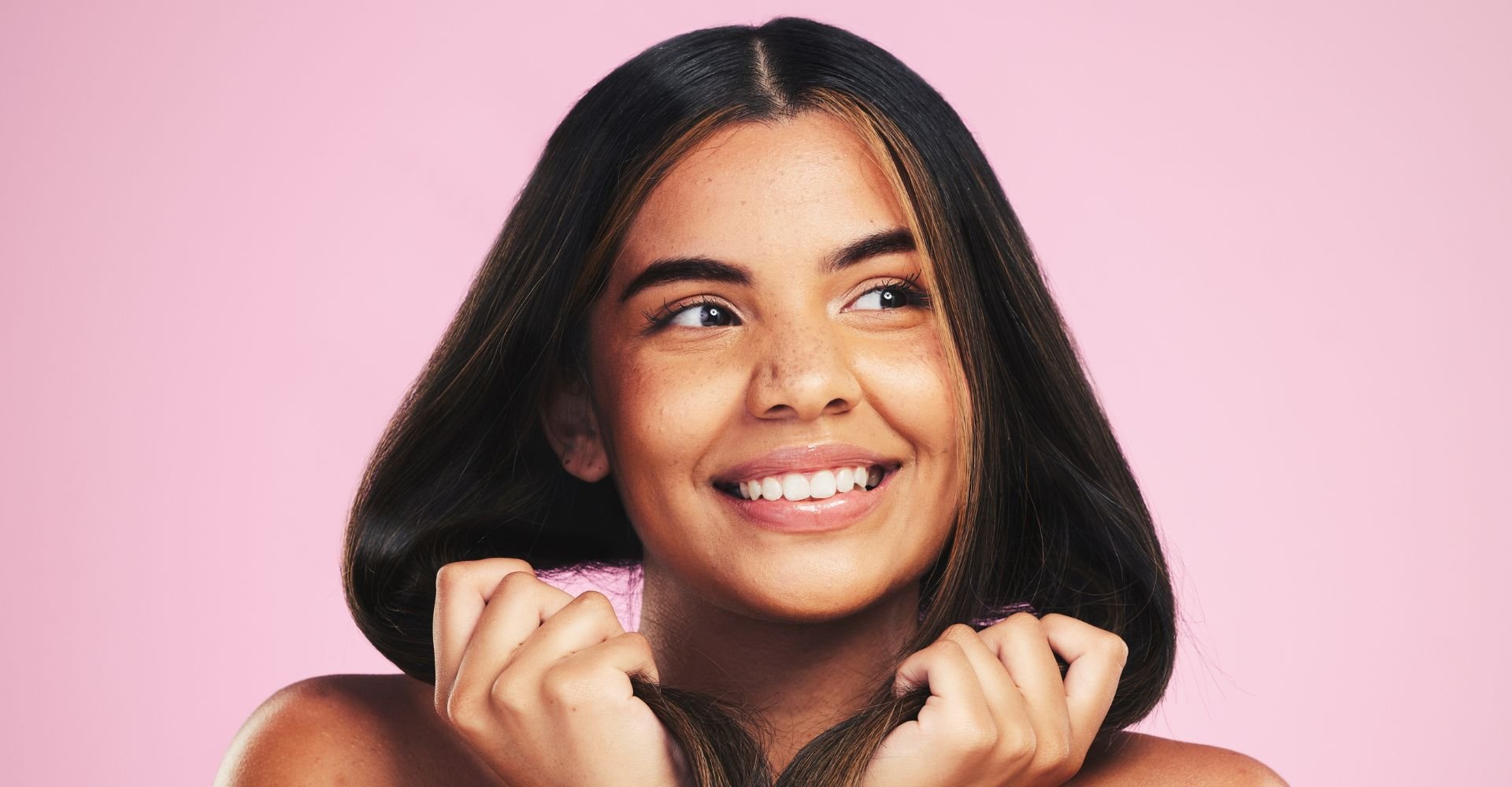 Mulher sorridente segurando seu cabelo com as mãos em fundo rosa.