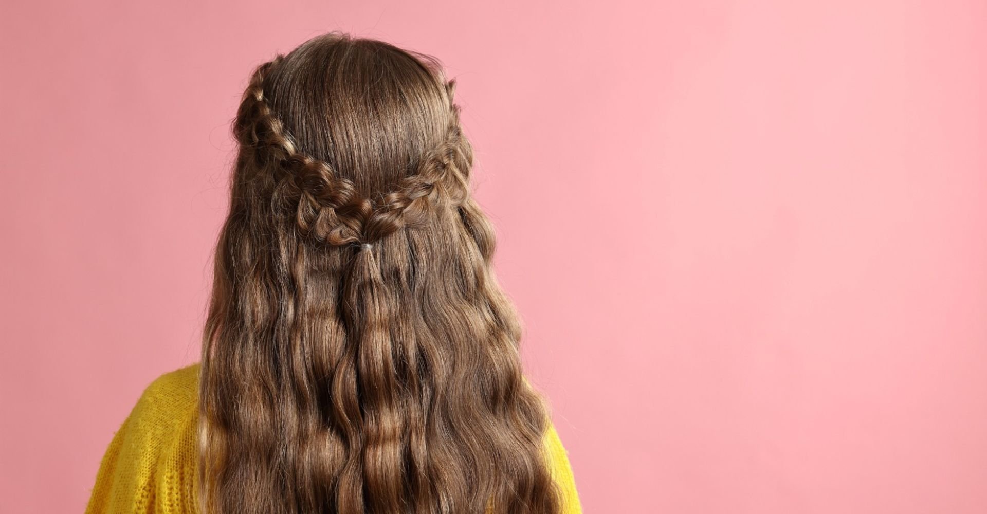 Mulher de costas com cabelo longo castanho ondulado, trança meia-cabeça presa ao centro, usando suéter amarelo. Fundo rosa
