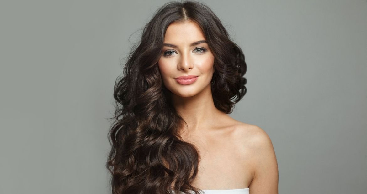 Mulher com olhos verdes, usando uma blusa branca e com cabelo castanho escuro ondulado.