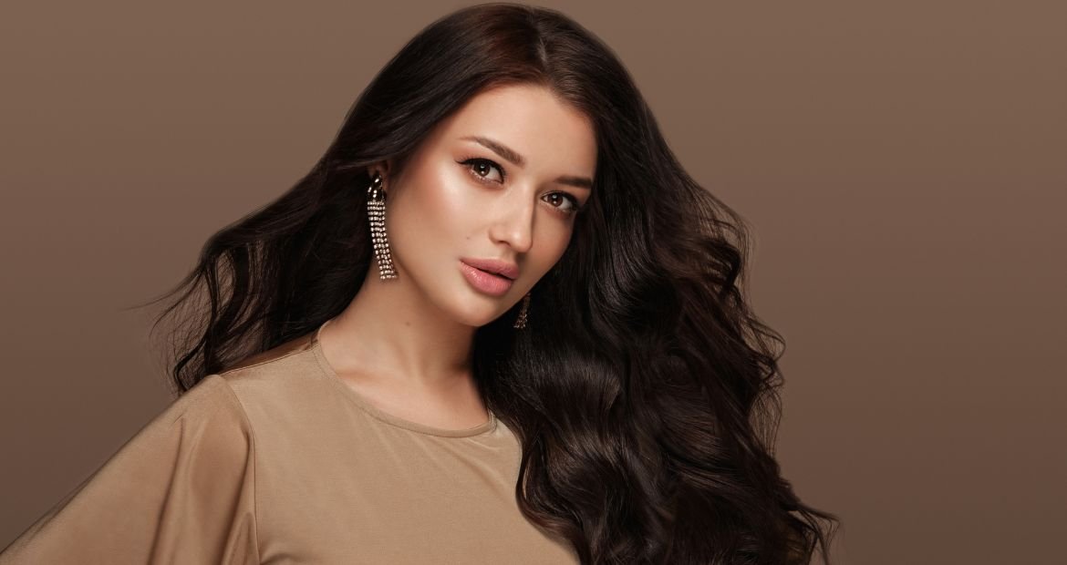 Mulher com cabelo castanho escuro liso, usando brincos longos e vestido bege. 