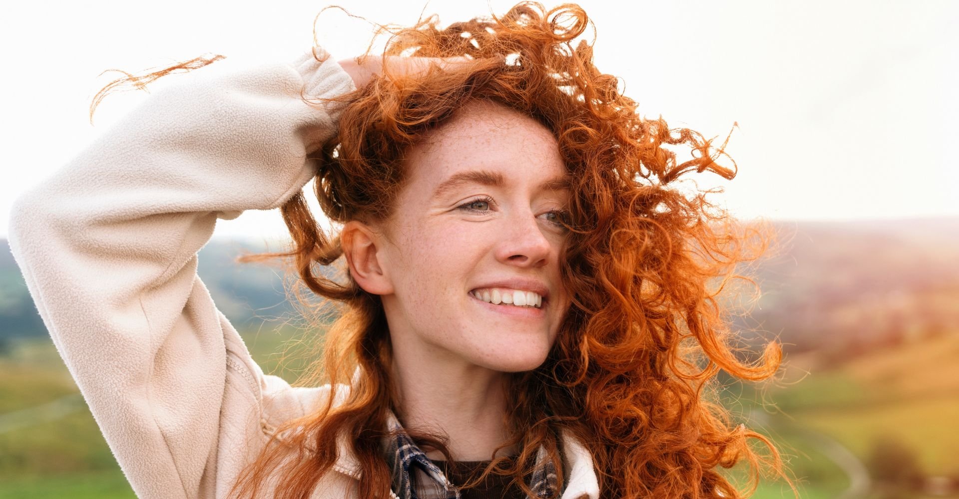 Menina sorridente com a mão no seu cabelo ruivo em uma paisagem verde atrás.