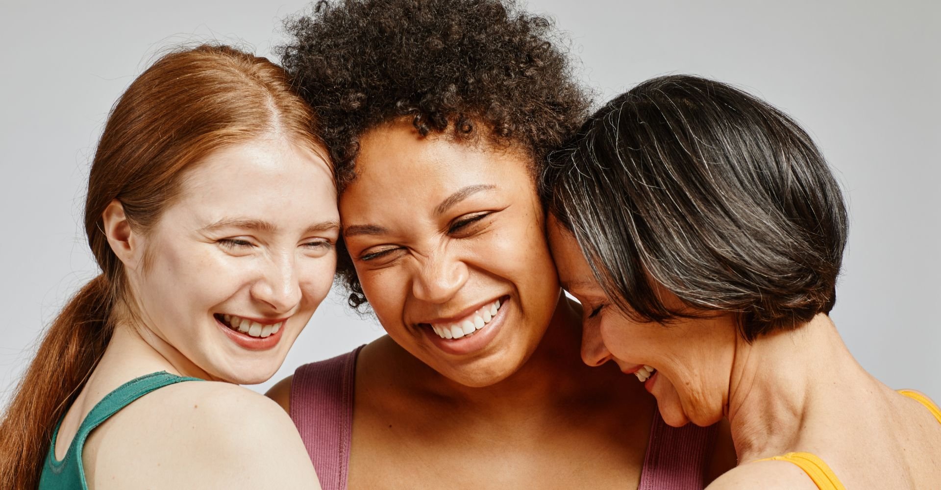Três mulheres com diferentes tons de pele e cores de cabelo sorrindo juntas, celebrando a diversidade e a beleza natural de cada uma.