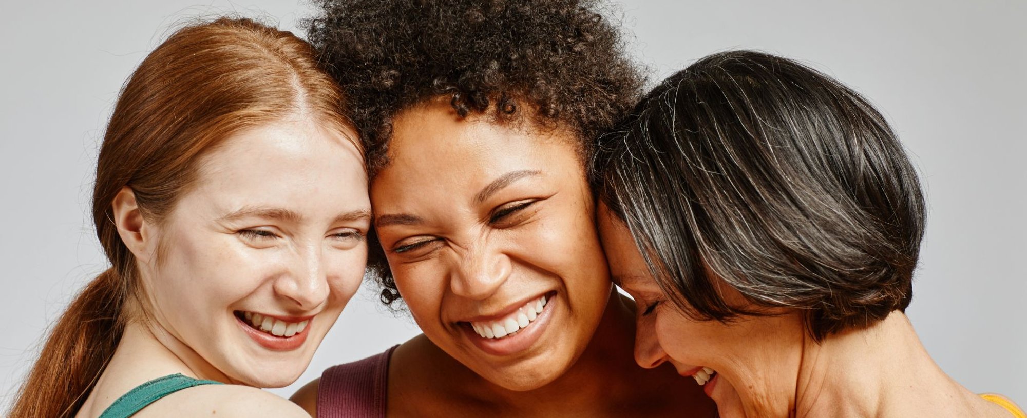 Três mulheres com diferentes tons de pele e cores de cabelo sorrindo juntas, celebrando a diversidade e a beleza natural de cada uma.