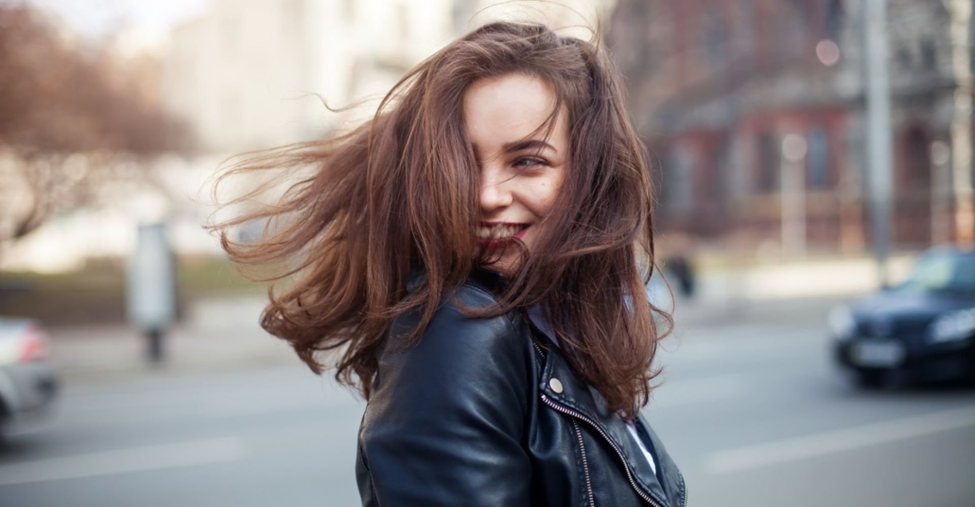 Mulher jovem com cabelo castanho médio ao vento sorrindo na rua, usando jaqueta de couro preta, com fios de tom quente e luminoso.