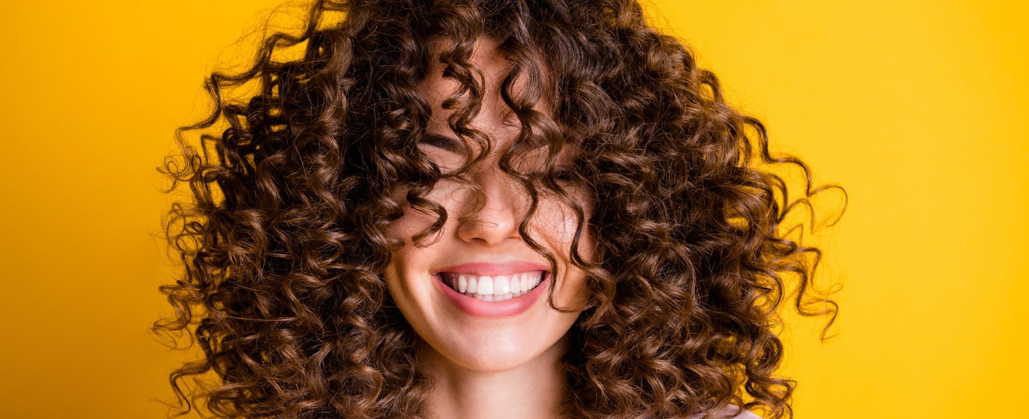 Mulher sorridente com cabelos castanhos cacheados e volumosos, usando camiseta clara em fundo amarelo, transmitindo alegria e autoestima.