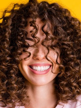 Mulher sorridente com cabelos castanhos cacheados e volumosos, usando camiseta clara em fundo amarelo, transmitindo alegria e autoestima.