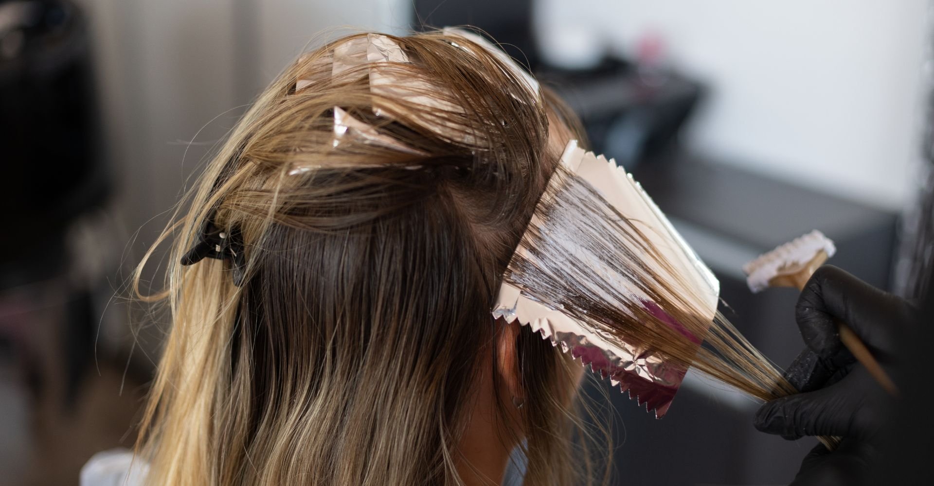 Mulher de cabelos loiros lisos recebendo aplicação de coloração nos fios com papel alumínio e pincel, realizada por profissional usando luvas pretas em salão de beleza.